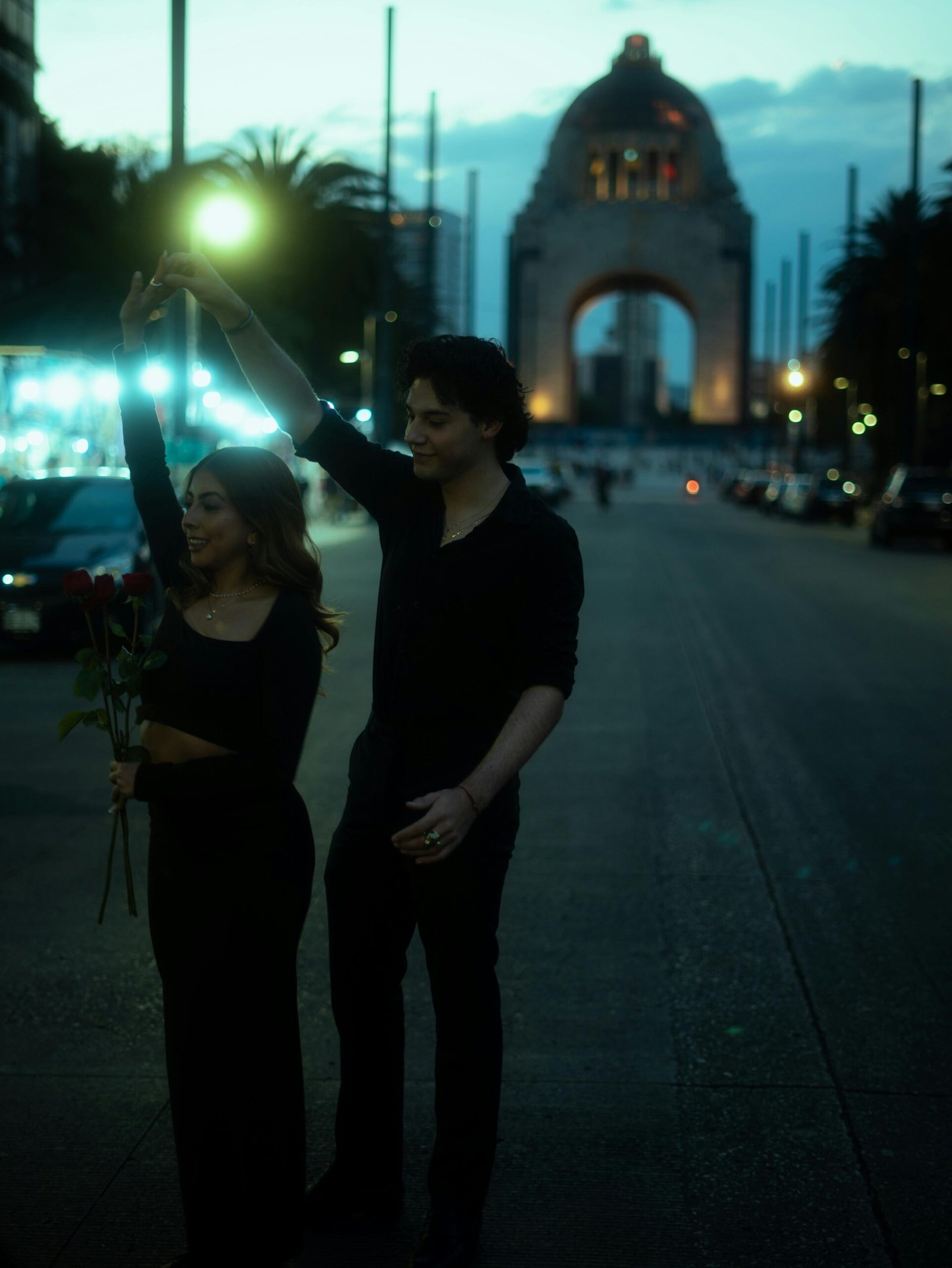 A couple shares a romantic moment with roses by a historic monument in Mexico City at night.